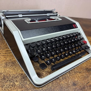 May include: A portable typewriter with a light gray frame and a black body. It features a QWERTY keyboard with black keys, a red key on the right side, and a metal carriage assembly at the back. The design includes a black top panel and a carriage return lever.