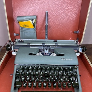 May include: A sage green Olympia De Luxe portable typewriter with a QWERTY keyboard, housed in a cream-colored carrying case with a textured terracotta interior. A small grey pouch containing a yellow cleaning cloth is attached to the case lid.