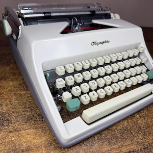 Peut inclure: Une machine à écrire Olympia dotée d'un corps couleur crème et d'une base grise. Le clavier comprend des touches circulaires blanches avec des lettres noires, accentuées par deux touches vert clair sur les côtés gauche et droit.