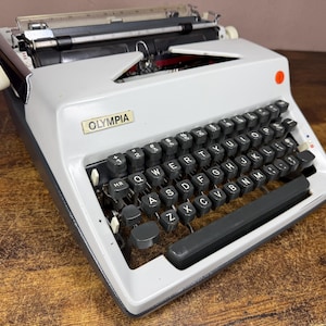 以下が含まれることがあります： 黒いキーを備えたQWERTYキーボード、金属製のキャリッジ、目立つブランド名プレートを特徴とするライトグレーのオリンピア製タイプライター。このデバイスには、スペースバー、さまざまなファンクションキー、および右上のケースにある赤い円形のインジケーターが含まれています。