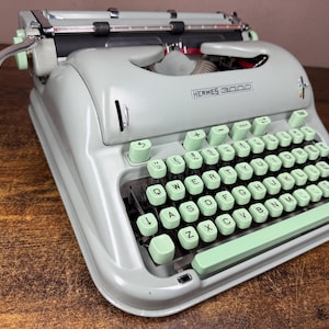 May include: A vintage Hermes 3000 typewriter in light grey with mint green keys. The keyboard has white letter keys and a mint green space bar. The typewriter features a silver carriage return lever and the brand name is visible.