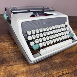 Puede incluir: Una máquina de escribir vintage blanca y gris con un teclado completo y detalles en color turquesa. La máquina de escribir está sobre una superficie de madera. La palabra "Royal" es visible en la parte delantera.