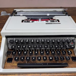 May include: A vintage Olivetti Lettera 32 typewriter in light grey. The typewriter has black keys with white lettering, a black space bar, and a silver carriage return lever. The machine sits on a wooden surface.