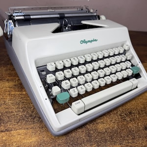 Puede incluir: Una máquina de escribir Olympia vintage en blanco y gris, con un teclado QWERTY completo y detalles en color turquesa. La palabra "Olympia" está impresa en turquesa en la parte delantera. La máquina está sobre una superficie de madera.