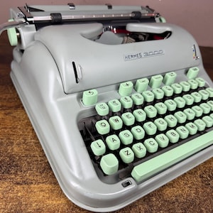 May include: A vintage Hermes 3000 typewriter featuring a light gray metal body and mint green keys. The QWERTY keyboard layout includes a long space bar, a carriage return lever on the left side, and the brand name printed on the front casing.