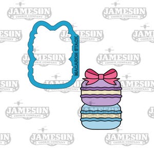 Puede incluir: Un cortador de galletas azul con forma de pila de macarons con un lazo rosa en la parte superior. El cortador está al lado de una pila de tres macarons con un lazo rosa en la parte superior. Los macarons son morados, azul claro y azules.