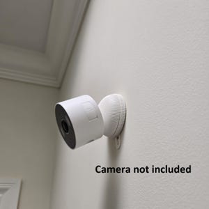 May include: A white security camera and its mounting bracket are attached to a white wall. The camera is cylindrical with a black lens and a white bracket. The text "Camera not included" is below the camera.