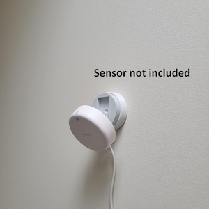 May include: A white, wall-mounted device with a circular base and a hinged, oval-shaped cover. A white cable extends from the device. The text "Sensor not included" is above the device.