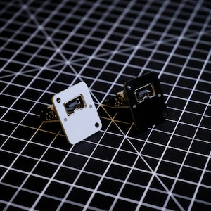 May include: Two small electronic components, one white and one black, are displayed. Each has a rectangular shape with rounded corners and a central opening. The white component is on the left, and the black one is on the right. Both are set against a black grid background.