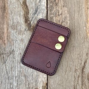 May include: A dark brown leather card holder with two brass button closures. The card holder has a stitched border and a small teardrop design. The card holder is rectangular and has two card slots.