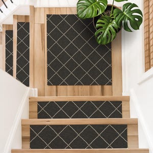May include: A set of dark charcoal gray stair treads and a matching rectangular floor mat, each featuring a white diamond grid pattern. The textile pieces are arranged on light-toned wooden stairs and a landing, with a green monstera plant.