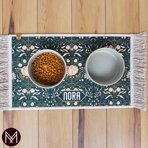May include: A green and gold floral patterned pet food mat with white fringe. The mat has two white ceramic bowls, one filled with brown kibble. The mat has the name "Nora" printed on it.