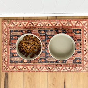 May include: Two white ceramic pet bowls on a red, blue, and cream patterned rug. One bowl contains dry dog food.