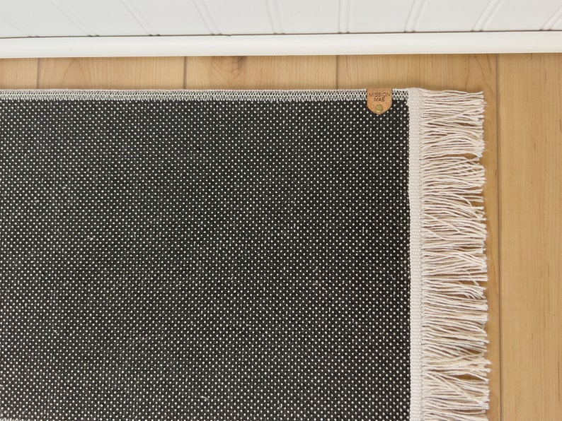 Puede incluir: Alfombra de patr&oacute;n blanco y negro con un borde de flecos blancos. La alfombra tiene una peque&ntilde;a etiqueta de cuero marr&oacute;n con el texto "Made in USA" en ella.