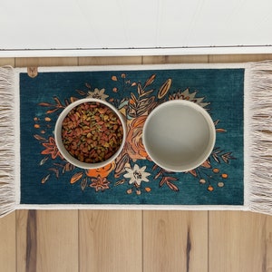 K&ouml;nnte beinhalten: Ein t&uuml;rkisfarbener Futterteppich f&uuml;r Haustiere mit floralem Design und zwei wei&szlig;en Keramikschalen. Eine Schale ist mit Trockenfutter gef&uuml;llt. Der Teppich hat einen Fransenrand und liegt auf einem Holzboden.