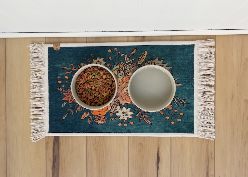 K&ouml;nnte beinhalten: Ein t&uuml;rkisfarbener Futterteppich f&uuml;r Haustiere mit floralem Design und zwei wei&szlig;en Keramikschalen. Eine Schale ist mit Trockenfutter gef&uuml;llt. Der Teppich hat einen Fransenrand und liegt auf einem Holzboden.