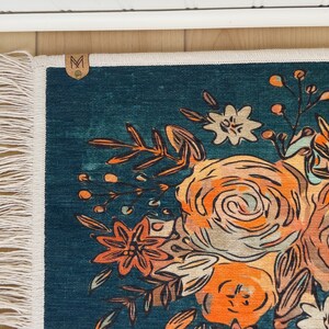 K&ouml;nnte beinhalten: Ein teal Teppich mit einem Blumendesign mit orangefarbenen, pfirsichfarbenen und wei&szlig;en Blumen. Der Teppich hat einen Fransenrand und ein kleines Lederetikett mit dem Buchstaben "M". Das Blumenmuster ist eine stilisierte Illustration.
