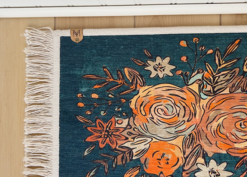 K&ouml;nnte beinhalten: Ein teal Teppich mit einem Blumendesign mit orangefarbenen, pfirsichfarbenen und wei&szlig;en Blumen. Der Teppich hat einen Fransenrand und ein kleines Lederetikett mit dem Buchstaben "M". Das Blumenmuster ist eine stilisierte Illustration.