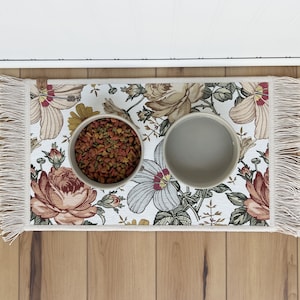 Puede incluir: Una alfombra blanca de comida para mascotas con flores y bordes con flecos y dos cuencos de cer&aacute;mica. Un cuenco est&aacute; lleno de comida para mascotas marr&oacute;n.
