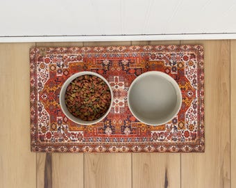 May include: A rectangular pet feeding mat featuring a detailed, intricate Persian-style rug pattern in shades of orange, red, navy blue, and white, with two round, light-coloured ceramic bowls placed on top, one filled with dry brown pet food.