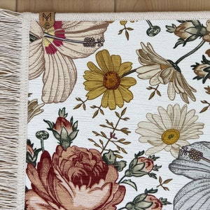 Puede incluir: Una alfombra blanca con un patr&oacute;n floral que presenta hibiscos, rosas y margaritas. La alfombra tiene un borde de flecos.