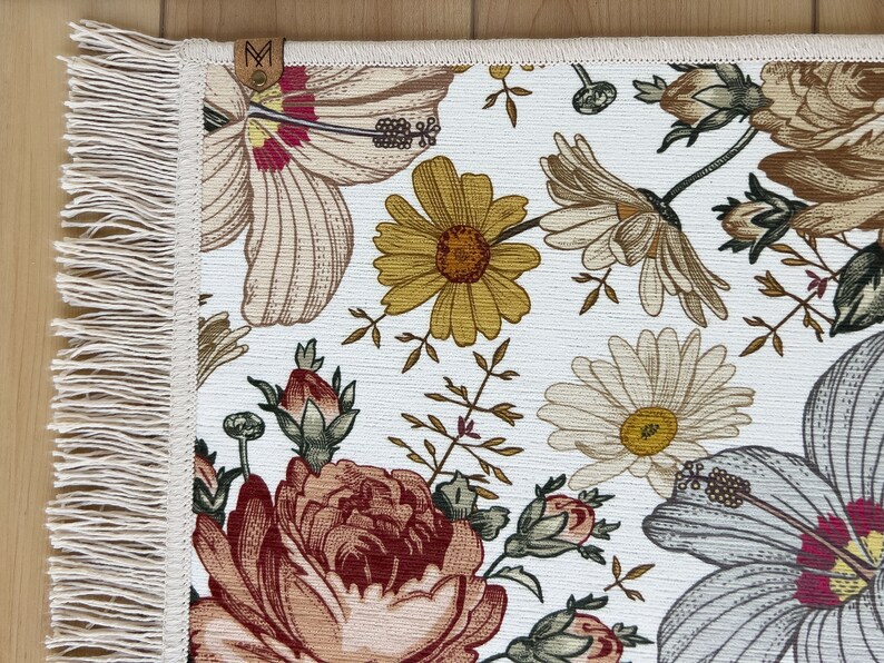 Puede incluir: Una alfombra blanca con un patr&oacute;n floral que presenta hibiscos, rosas y margaritas. La alfombra tiene un borde de flecos.