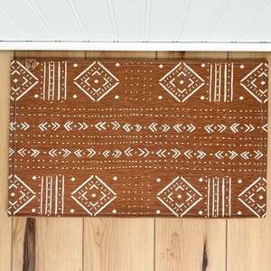 May include: A brown doormat with a white geometric pattern. The pattern includes diamonds, arrows, and lines. The doormat has a "M" in the top left corner.