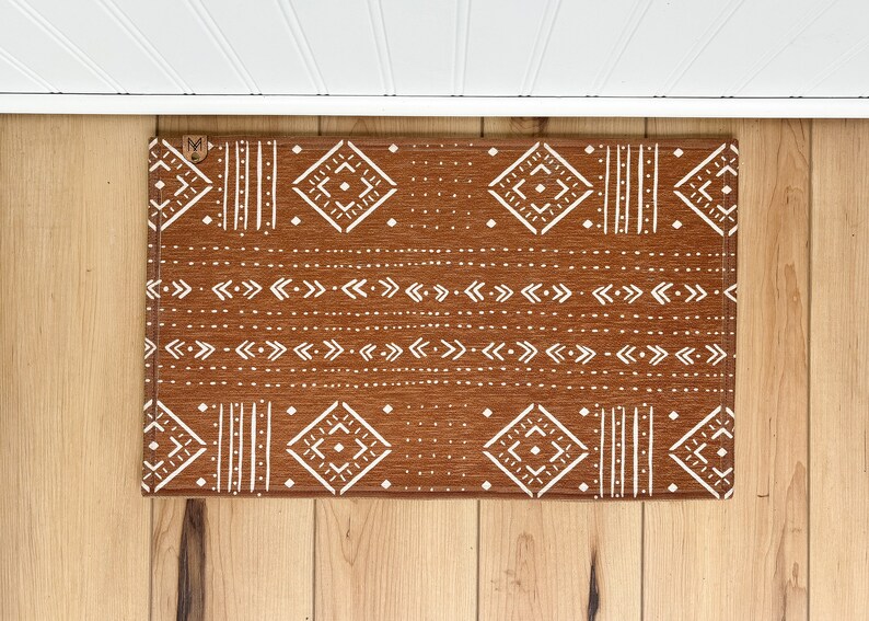 May include: A brown doormat with a white geometric pattern. The pattern includes diamonds, arrows, and lines. The doormat has a "M" in the top left corner.