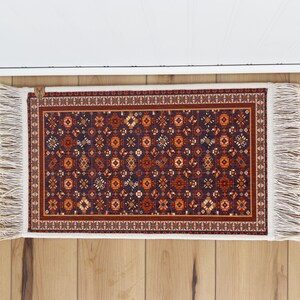 May include: A small, rectangular rug with a dark blue background and a repeating pattern of orange, red, and white geometric shapes. The rug has white fringe on all sides.