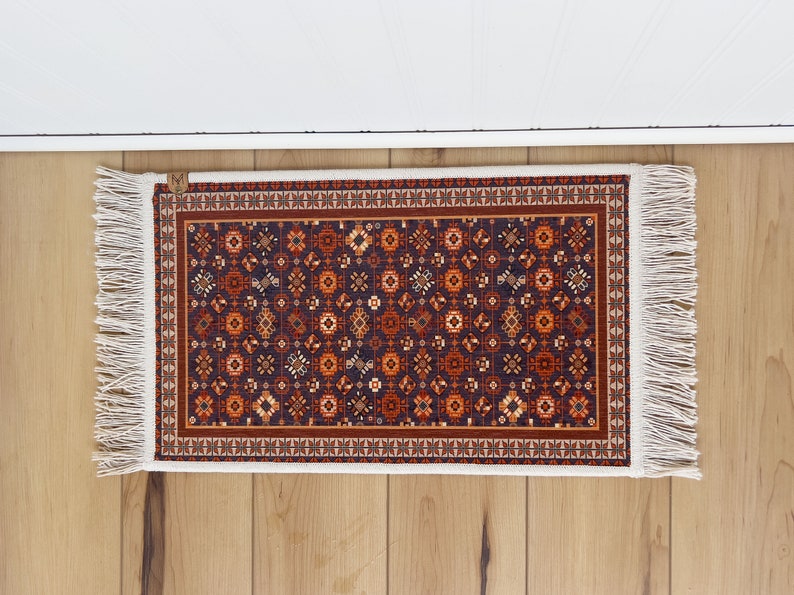 May include: A small, rectangular rug with a dark blue background and a repeating pattern of orange, red, and white geometric shapes. The rug has white fringe on all sides.