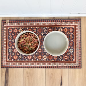 May include: A brown and orange patterned dog bowl mat with two white ceramic bowls, one filled with dog food and the other empty.