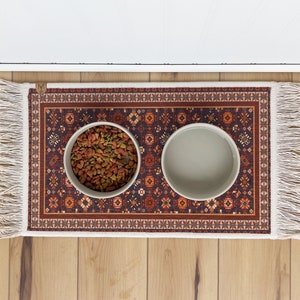 May include: A red, orange, and blue patterned rug with fringe on the edges. Two white ceramic bowls are on the rug, one filled with pet food.
