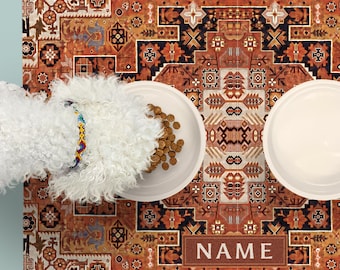 May include: A rectangular pet feeding mat featuring an intricate orange, brown, and black geometric pattern with a central rectangular box containing the text NAME. Two white bowls sit on the mat, one filled with brown kibble.