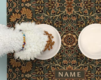 May include: A rectangular pet feeding mat featuring a dark green and brown floral pattern with a central rectangular box containing the word NAME. Two white bowls sit on the mat, one filled with brown kibble and one empty.