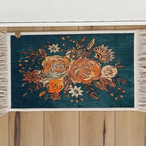 K&ouml;nnte beinhalten: Rechteckiger tealfarbener Teppich mit floralem Design. Der Teppich zeigt einen Strau&szlig; aus orangefarbenen und beigefarbenen Blumen mit gr&uuml;nen Bl&auml;ttern. Der Teppich hat einen wei&szlig;en Fransenrand und liegt auf einer Holzoberfl&auml;che.