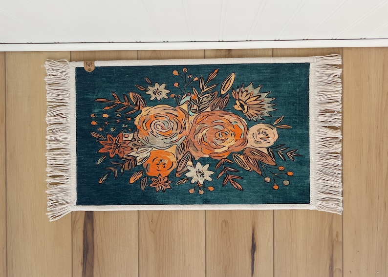 K&ouml;nnte beinhalten: Rechteckiger tealfarbener Teppich mit floralem Design. Der Teppich zeigt einen Strau&szlig; aus orangefarbenen und beigefarbenen Blumen mit gr&uuml;nen Bl&auml;ttern. Der Teppich hat einen wei&szlig;en Fransenrand und liegt auf einer Holzoberfl&auml;che.