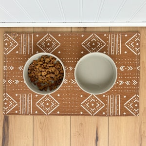 May include: A brown and white patterned pet food mat with two white ceramic bowls. One bowl is filled with brown dog food.