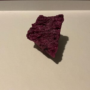 May include: A deep magenta, irregularly shaped mineral specimen with a rough, porous, and crystalline texture. The object features sharp, jagged edges and a dense, uneven surface structure that catches light, creating small shadows across its form.