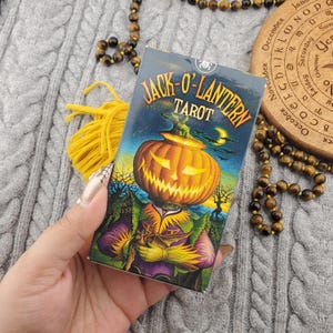 Puede incluir: Una baraja de cartas de tarot titulada "Jack-O-Lantern Tarot" con una figura con cabeza de calabaza. La ilustración es colorida con un tema de Halloween. La baraja se sostiene en una mano, con una borla amarilla y joyas de cuentas en el fondo.