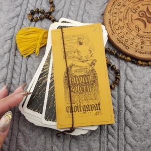 May include: A tarot card deck with a mustard yellow cover featuring text and an illustration. The deck rests on a grey knitted surface, with a yellow tassel and a wooden circular object with text. A brown beaded necklace is also present.