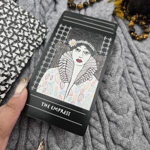 K&ouml;nnte beinhalten: Eine schwarze Tarotkarte mit einer Illustration einer Frau mit Blumenkrone und gemustertem Mantel. Die Karte tr&auml;gt die Aufschrift "THE EMPRESS". Die Karte wird in einer Hand gehalten.