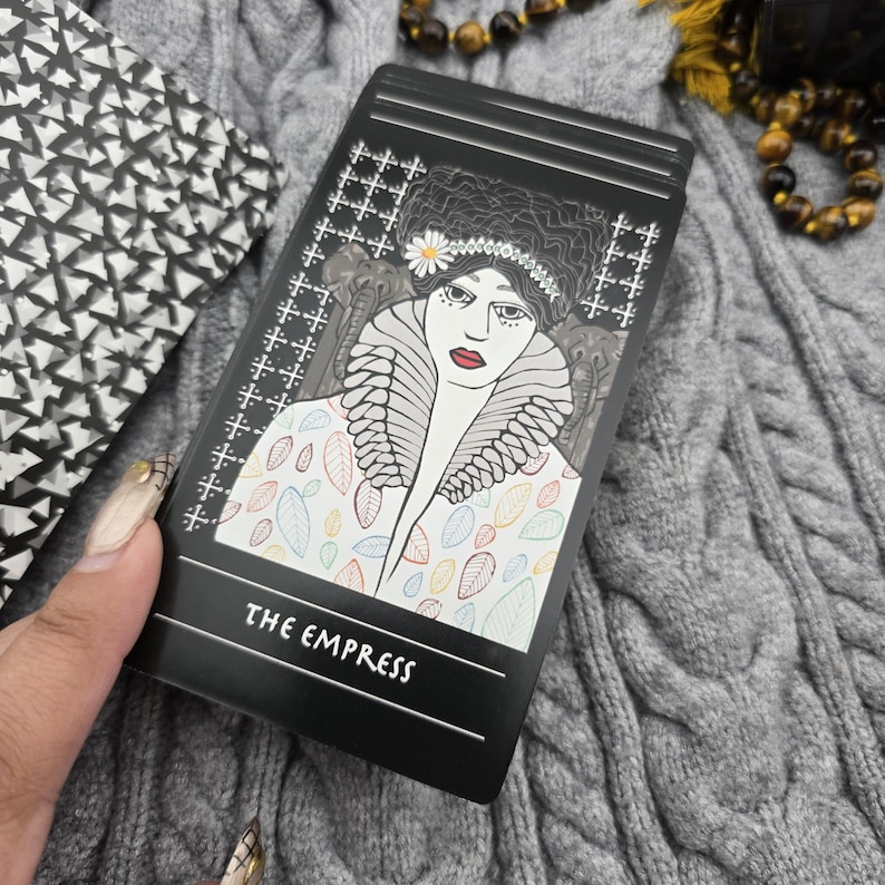 K&ouml;nnte beinhalten: Eine schwarze Tarotkarte mit einer Illustration einer Frau mit Blumenkrone und gemustertem Mantel. Die Karte tr&auml;gt die Aufschrift "THE EMPRESS". Die Karte wird in einer Hand gehalten.