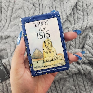 Könnte beinhalten: Eine blaue Tarotkartenbox mit den Worten "TAROT der ISIS" in Weiß. Die Box zeigt eine Illustration der Sphinx und einer Pyramide. Die Box hat auch Text auf Deutsch. Die Box enthält 36 Karten.