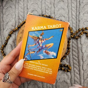 May include: A rectangular Karma Tarot deck box with an orange background and a central illustration of a figure with wings and a sword against a blue sky. The text reads Karma Tarot by Birgit Boline Erfurt, the 78-card Karma Tarot deck is a fascinating blend of colour, motion and phantasmagoric art, complete with instructions.