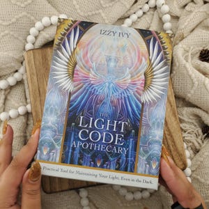 Op de afbeelding: Een rechthoekig boek getiteld The Light Code Apothecary door Izzy Ivy, met een kleurrijke, symmetrische illustratie van een gevleugelde figuur in blauwe, paarse en gouden tinten, met de ondertitel A Practical Tool for Maintaining Your Light.