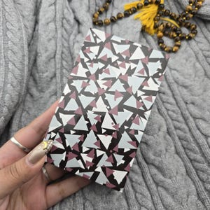 May include: A rectangular box featuring a geometric design of white, grey, and maroon triangles on a black backdrop. The box is held in a hand with silver rings. The background is a grey cable-knit jumper.