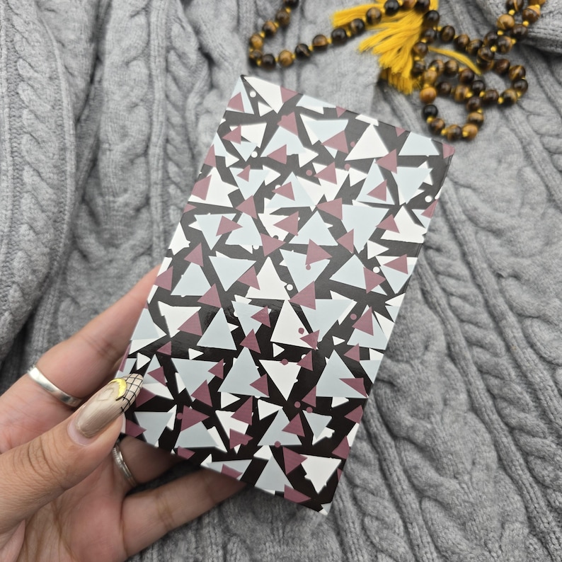 K&ouml;nnte beinhalten: Eine rechteckige Schachtel mit einem geometrischen Muster aus wei&szlig;en, grauen und weinroten Dreiecken auf schwarzem Hintergrund. Die Schachtel wird in einer Hand mit silbernen Ringen gehalten. Der Hintergrund ist ein grauer Zopfstrickpullover.