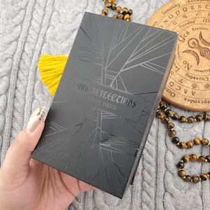 Op de afbeelding: Een zwarte tarotdeckdoos met de woorden "DARK REFLECTIONS" en "TAROT DECK" in een gestileerd lettertype. De doos heeft een geometrisch ontwerp en wordt in een hand gehouden. Een gele kwast en een kralenketting zijn ook zichtbaar.