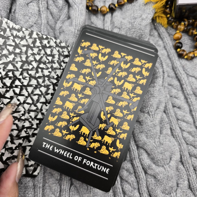 K&ouml;nnte beinhalten: Ein Tarotkartenspiel mit schwarzem Hintergrund und goldenen und grauen Illustrationen. Die im Fokus stehende Karte ist "Das Gl&uuml;cksrad" mit einem Raddesign und Tierfiguren. Der Text "THE WHEEL OF FORTUNE" befindet sich unten.