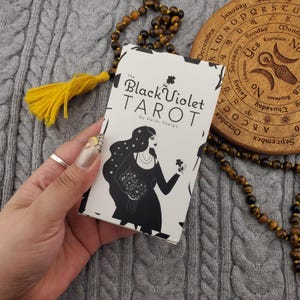 Könnte beinhalten: Ein Tarotkartenspiel mit dem Titel "The Black Violet Tarot" von Heidi Phelps. Die Schachtel zeigt eine Schwarz-Weiß-Illustration einer Frau, die Blumen hält. Ein gelber Quastenanhänger und eine Perlenkette sind im Hintergrund zu sehen.
